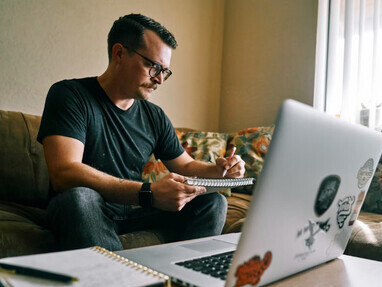 In the soft light of early morning, a man in a black crew-neck t-shirt relaxes on the couch, deeply absorbed in his drawing