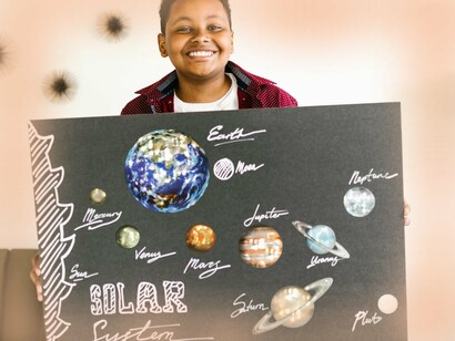 Estudante posa orgulhoso com sua montagem do sistema solar. Ao conectar o aprendizado ao seu próprio repertório, o aluno se torna mais capaz de buscar e aplicar o conhecimento de maneira independente