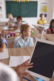 In un'aula scolastica, un'insegnante in primo piano tiene in mano un tablet, i suoi studenti sono seduti ai banchi
